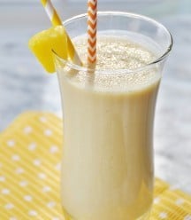 A glass with a yellow smoothie with a pineapple wedge on the edge of the glass and an orange and white straw on a yellow and white polka dot napkin.