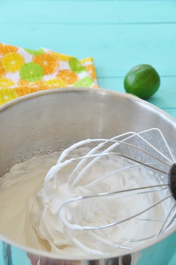 Vegan Meringue in a silver mixing bowl with a whisk attachment and meringue clinging to it on a turquoise board with a lime and an assorted citrus print towel