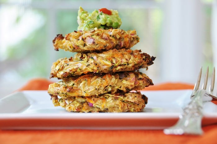 A stack of four Vegan Oil-Free Zucchini & Carrot Fritters with a dollop of guacamole on top on a square whit plate with a silver fork on an orange napkin
