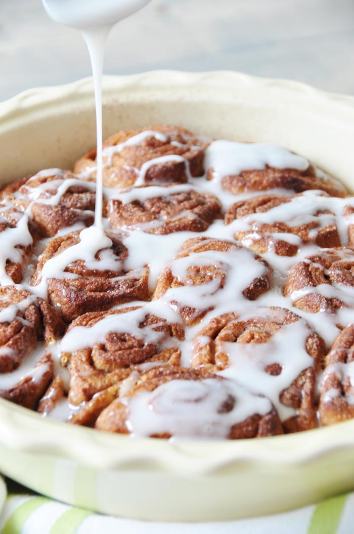 Homemade Vegan Cinnamon Rolls in a round yellow pie pan being drizzled with glaze