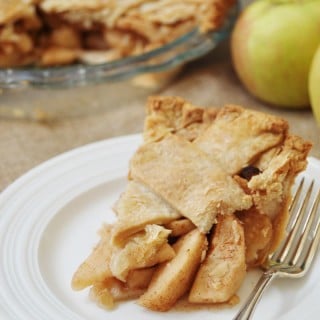 A slice of Homemade Apple Cinnamon Pie with a Flaky Vegan Pie Crust! on a white plate with a silver fork and the whole pie and an apple in the background