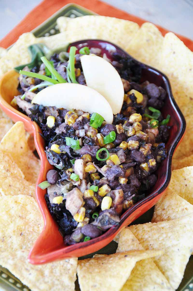 Better Beans Cuban Black Bean Dip in a red pepper shaped dish with two apple wedges and a sprig of rosemary on top and corn chips surrounding the bowl