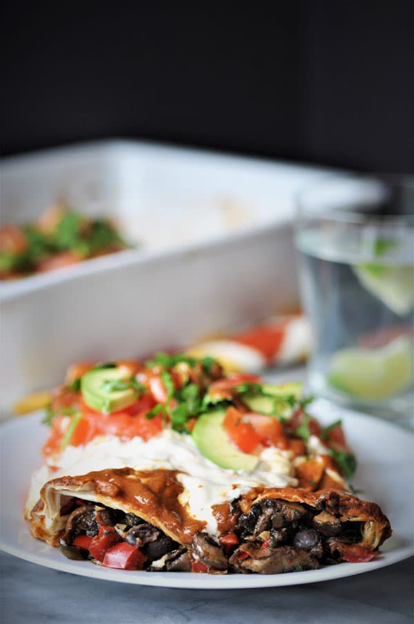 vegan black bean & mushroom enchiladas on a white plate with cashew cream sauce and sliced avocados and chopped cilantro.