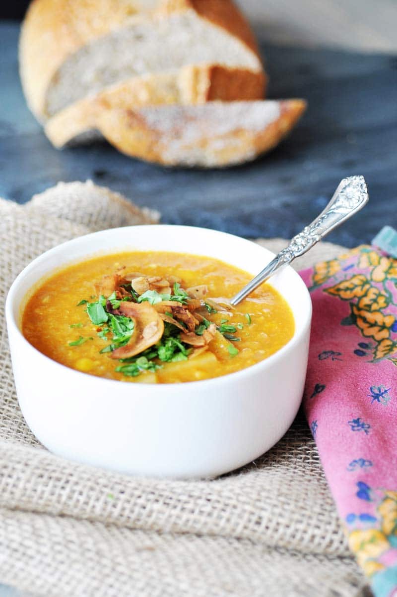 Corn chowder in a white bowl with a silver spoon sticking out of it and a loaf of sliced bread in the background.