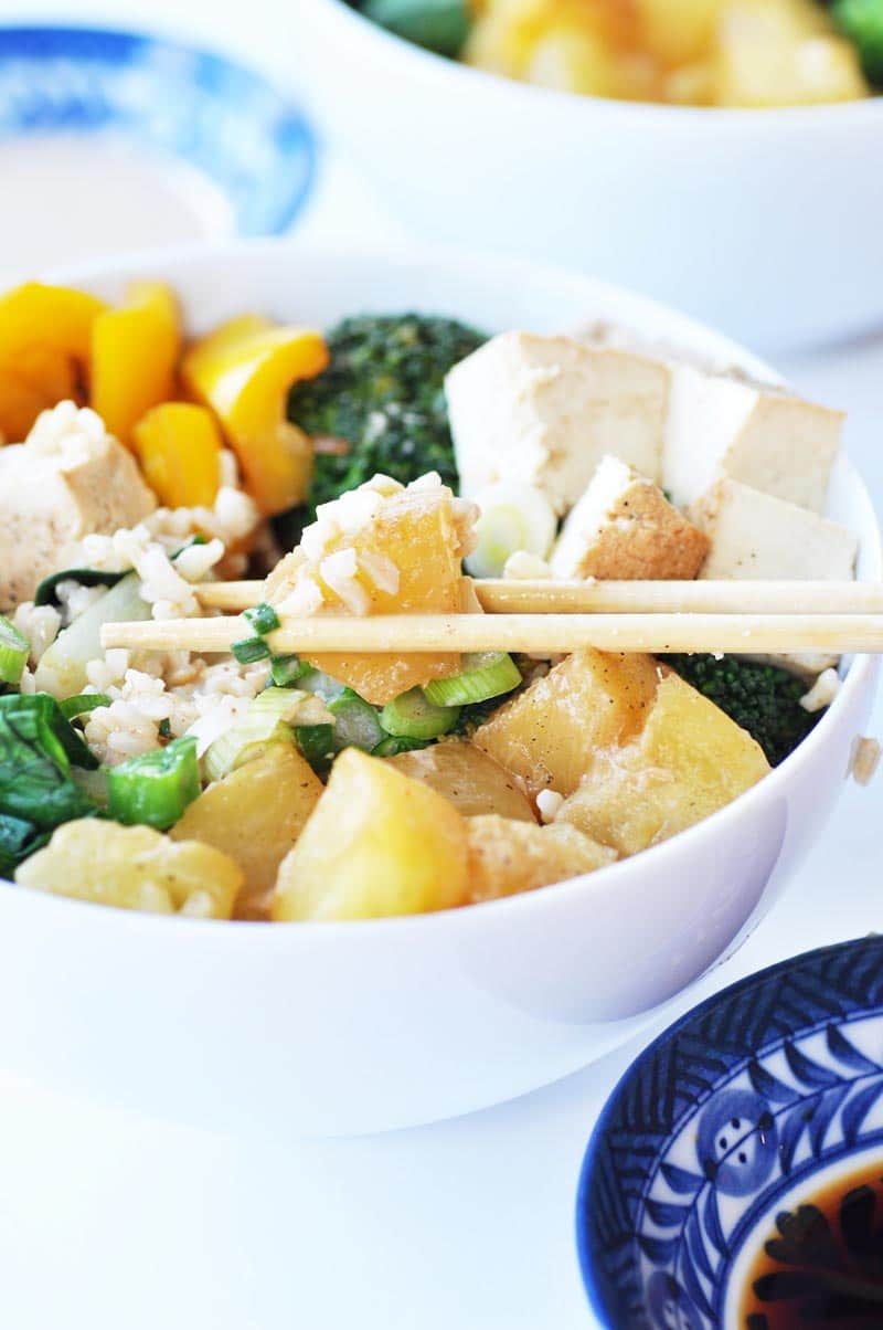 A white bowl with rice, pineapple, and broccoli and chopsticks picking up a piece of pineapple