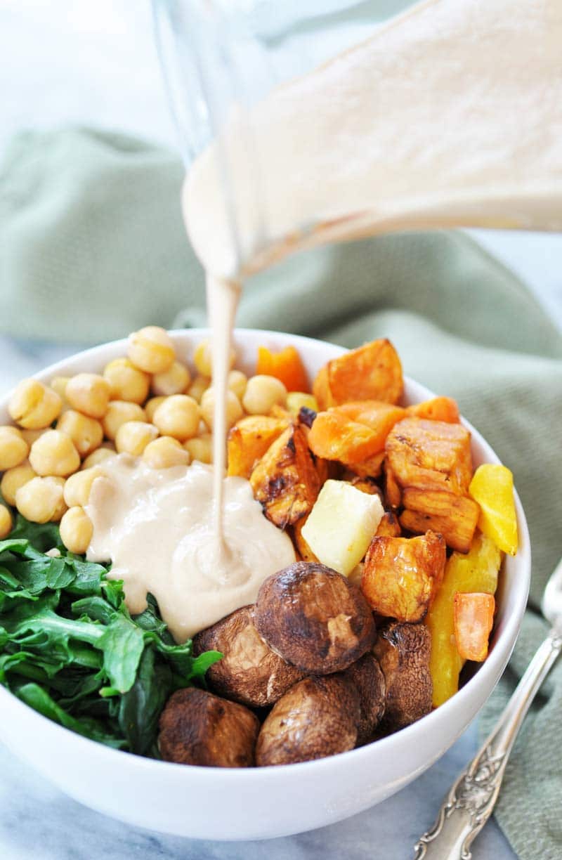A white bowl filled with roasted vegetables with a glass pitcher of dressing being poured over it