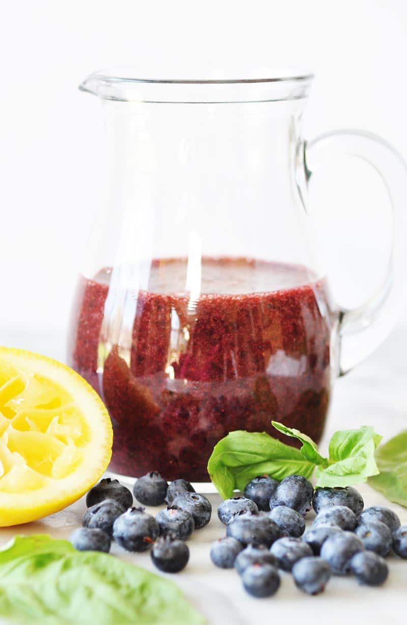 Blueberry Basil Salad Dressing in a clear glass pitcher with a lemon slice, fresh blueberries, and basil around it on a white surface