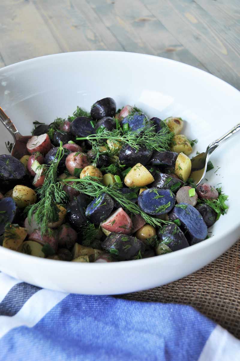 Tri-colored potato salad with dill in a white bowl with silver serving utensils and a blue and white napkin next to the bowl. 