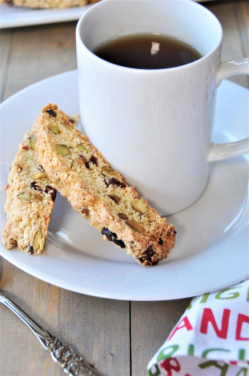 2 cranberry biscotti cookies on a saucer with a white mug of coffee and a red and green napkin next to the saucer.