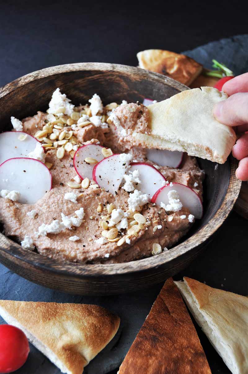Tahini and roasted beet hummus in a wood bowl with pita chips around it and a hand dipping a chip into the hummus