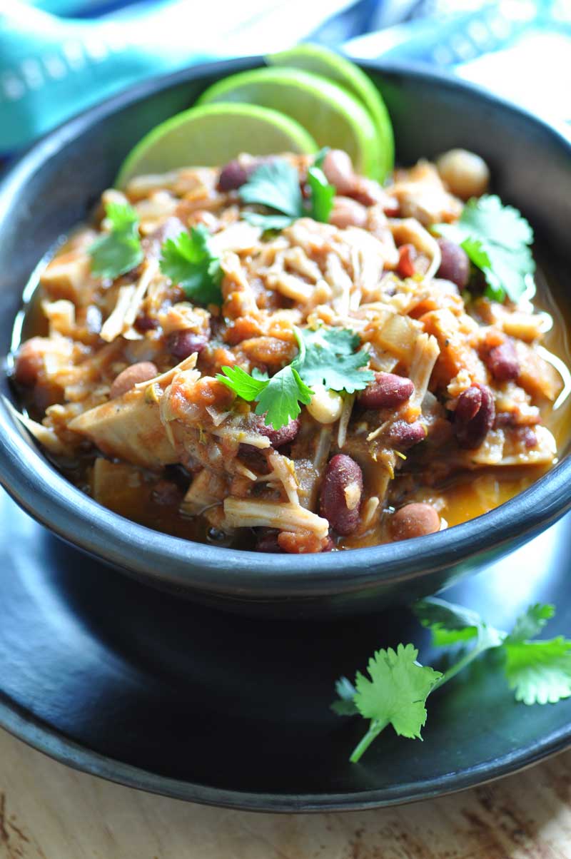 Jamaican Jerk Chik'n Chili in a black bowl on a balck plate with three lime wedges and sprigs of cilantro