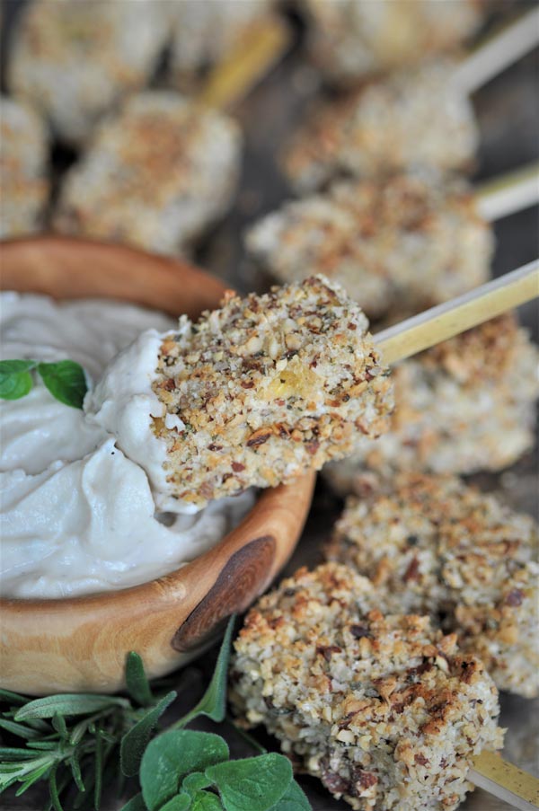 A skewer with a crusted almond tofu bite being dipped into lemon ginger dip in a wood bowl.