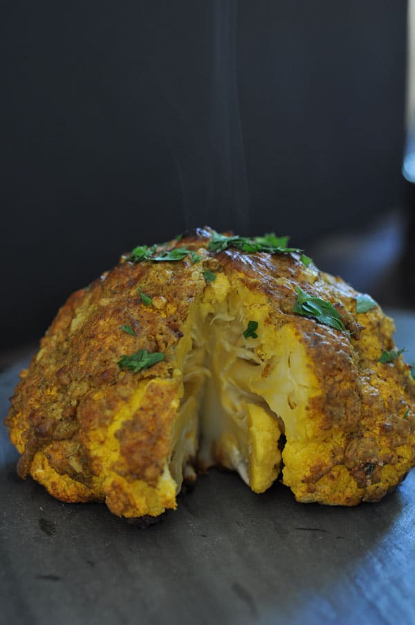 A steaming whole roasted cauliflower with a slice removed from it.