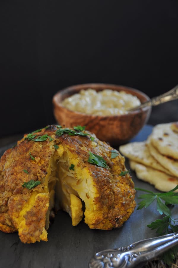 A whole roasted cauliflower on a slate board with parsley sprinkled over it and a wood bowl of sauce and naan in the background.