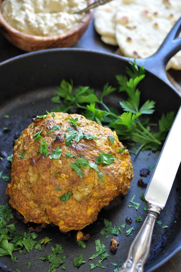 A whole roasted cauliflower in an iron skilled with parsley and a silver knife resting on the edge of the pan and a wood bowl of suace and naan in the background