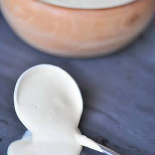The ultimate vegan cheese sauce dripping off of silver spoon on a slate surface with a wood bowl full of the cheese.