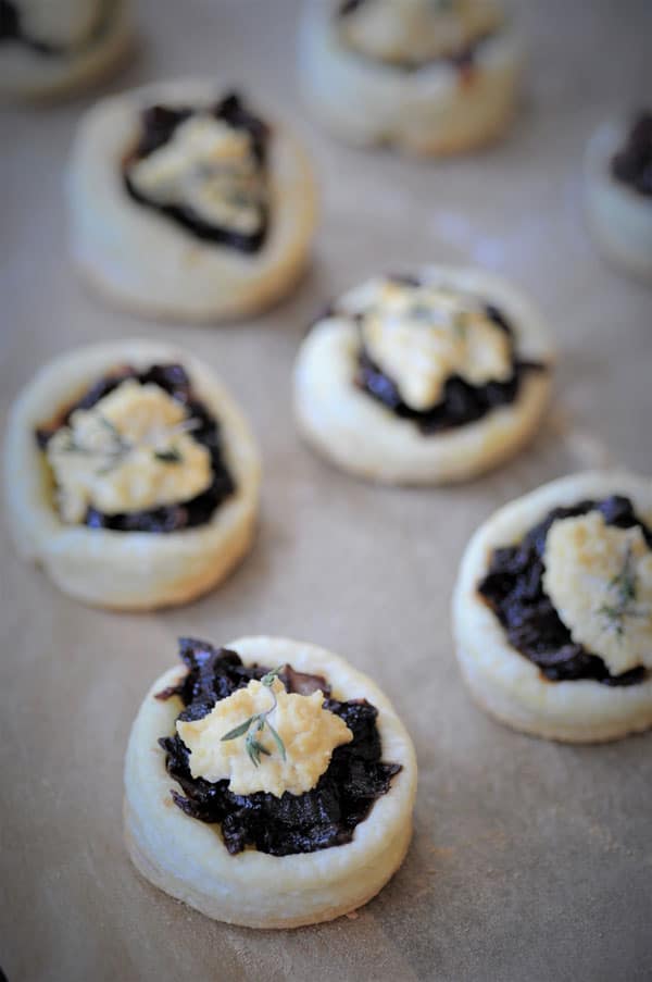 6 puff pastries with onion confit and a dollop of vegan ricotta and a sprig of thyme on parchment.
