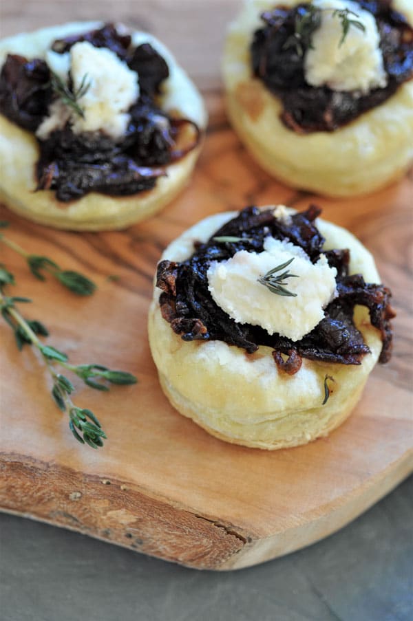 A close up of an onion confit tartlet on a wood board with a sprig of thyme.
