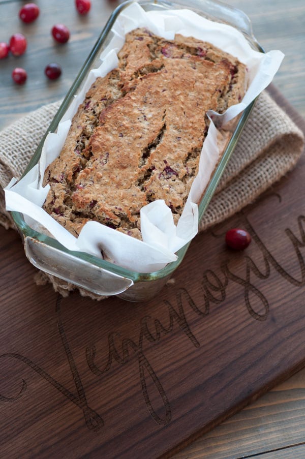 Vegan cranberry orange quick bread in a glass bread pan with parchment paper, sitting on a burlap cloth on a wood board.