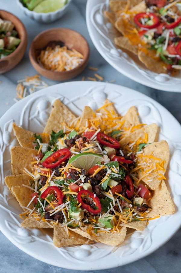 Loaded vegan nachos in two white bowls with 2 wood bowls filled with vegan shredded cheese and salsa, and a white bowl of lime wedges