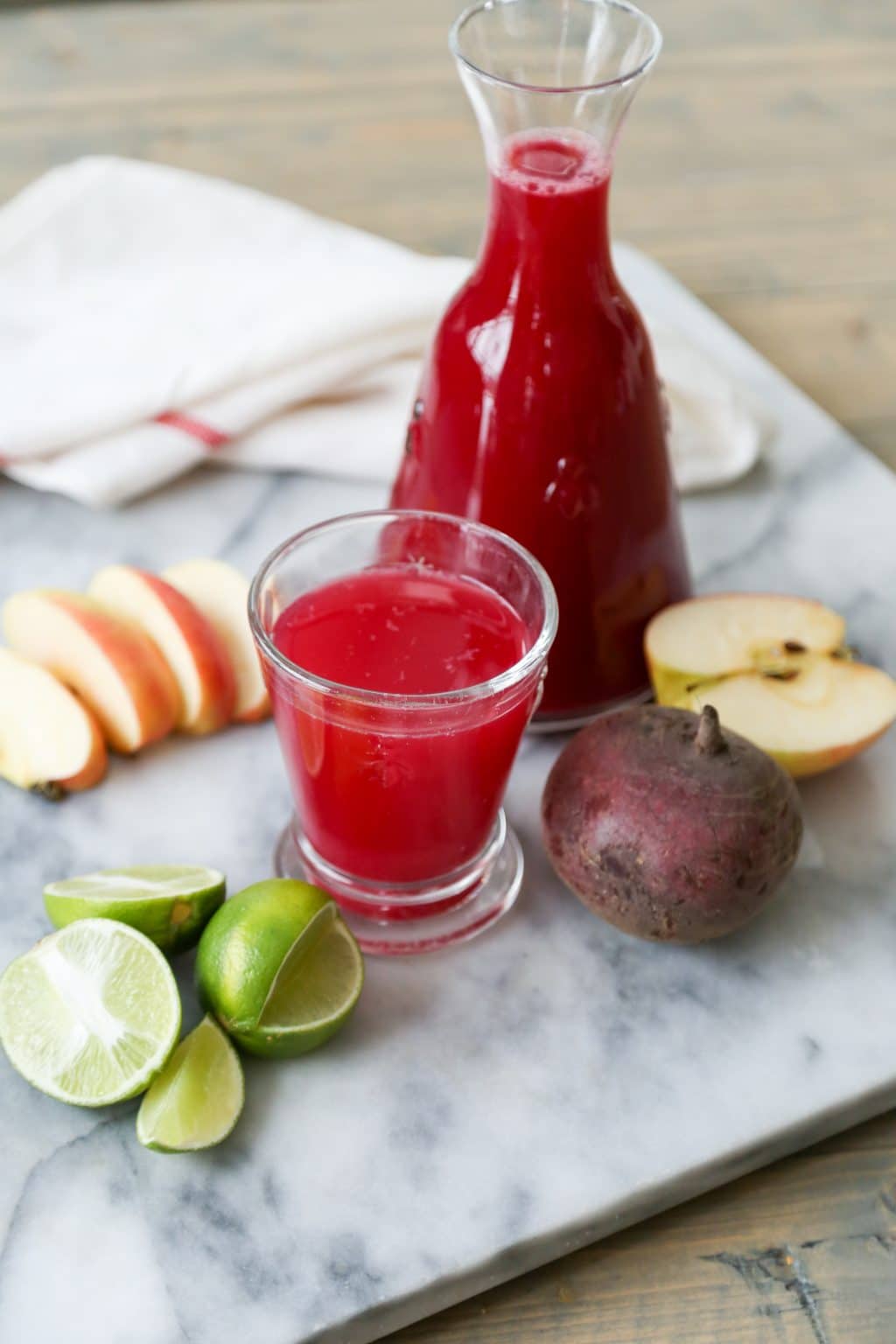 Beet, apple and lime juice in a carafe and a short glass on a marble slap with sliced apples, a beet and sliced limes scattered around.