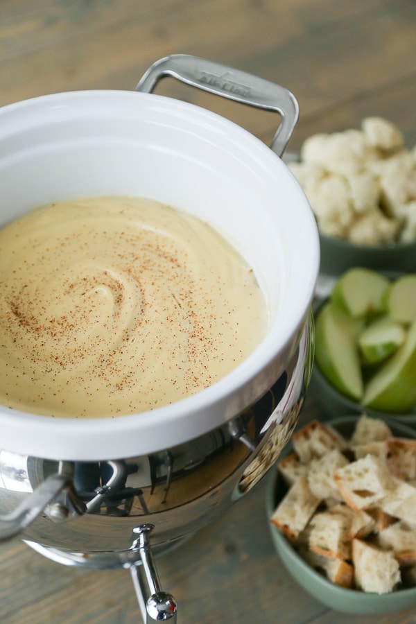 Vegan Fondue in a white fondue pot with bowls of bread, cauliflower, and apple slices on the side for dipping