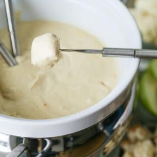 A white ceramic fondue pot filled with golden vegan cheese with a fondue fork dipping a piece of bread into the cheese
