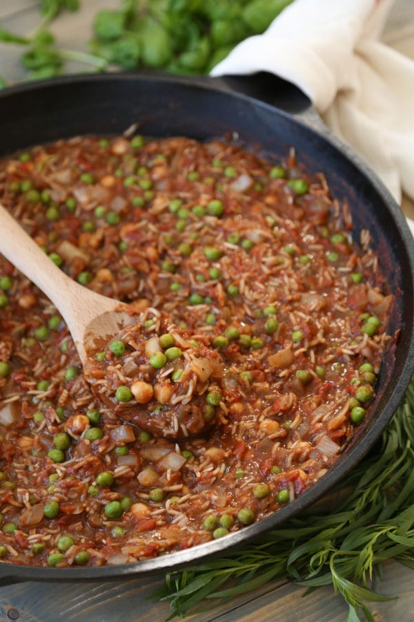 Indian Spiced Tomato & Rice Stew with chickpeas and green peas in a cast iron skillet with a wooden spoon stirring the stew. A white towel is wrapped around the handle and fresh tarragon is next to the pan