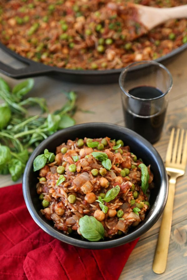 Indian spiced stew in a black bowl garnished with basil leaves with a gold fork, a red napkin, a glass of red wine, fresh herbs, and a cast iron pan of the stew next to the bowl