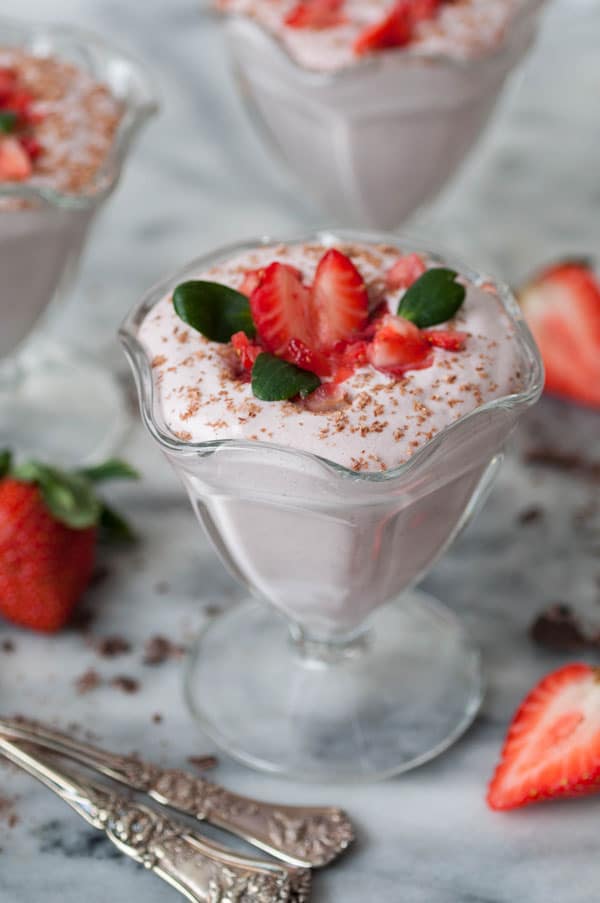 Three dishes of vegan strawberry mousse on a marble counter, garnished with chopped strawberries and shaved dark chocolate