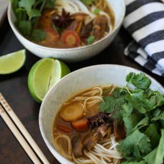 Two bowls of shiitake pho in white speckled bowls garnished with fresh cilantro and basil with chopsticks and lime wedges and a blue and white striped towel next to them