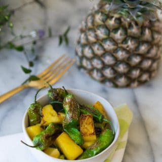 Shishito Peppers with pineapple in a white bowl with a gold fork and half a pineapple in the background