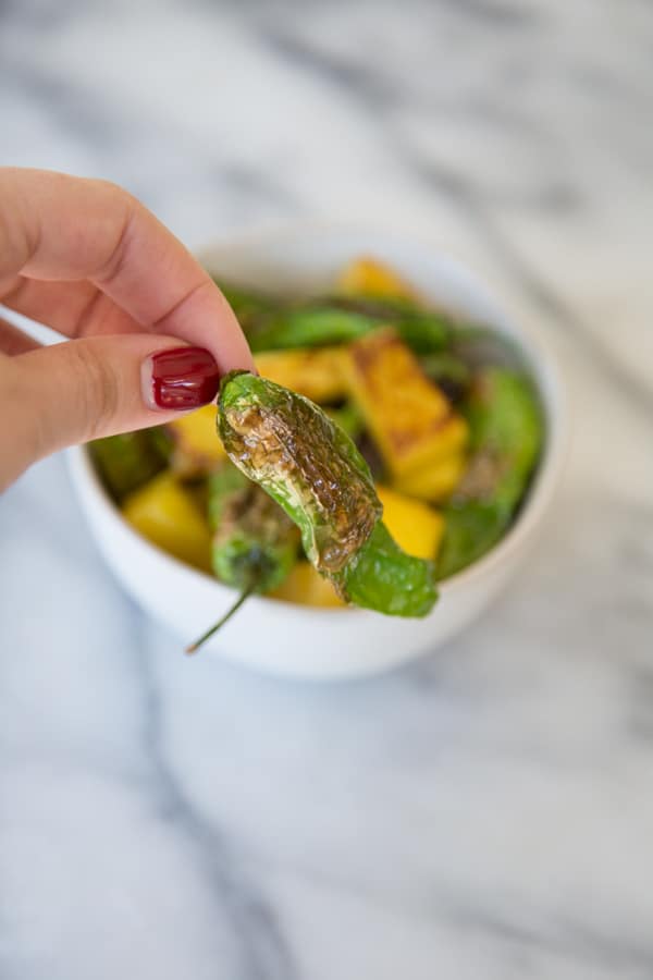 Shishito peppers being held in front of a white bowl filled with peppers and pineapple on a marble board