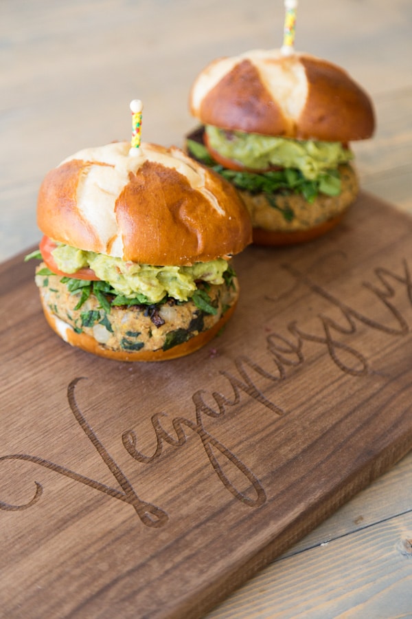 Two Spinach Burgers with shredded spinach, tomato slices, mashed avocado and buns with yellow and red toothpicks stuck on top of the bun. They're on a brown wood board with the word "Veganosity" etched into it.