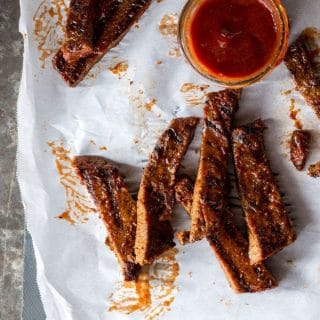 Slices of "Honey" BBQ BBQ ribz scattered on parchment paper with a dish of BBQ sauce in the upper right corner