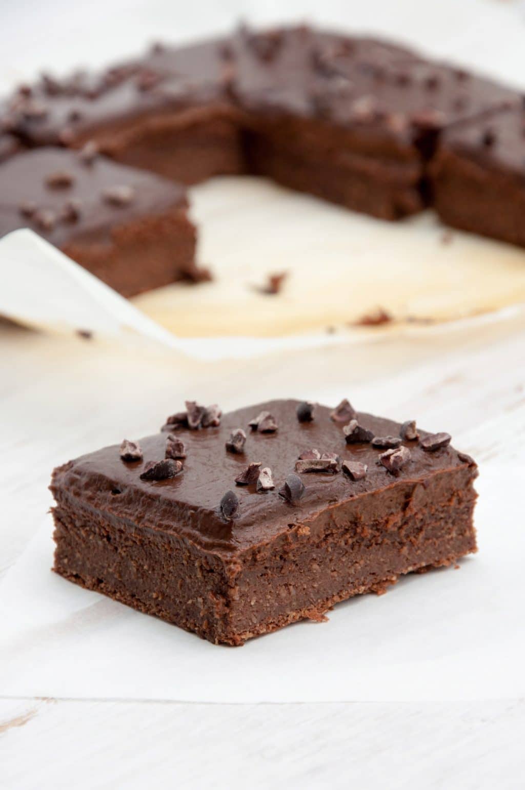 A Blender Brownie with frosting on a white surface with a pan of brownies in the background