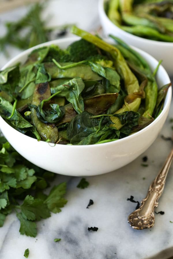 A bowl of pan roasted broccoli, snow peas, spinach, and green onions with another bowl in the background, with a silver fork and extra greens on a marble surface