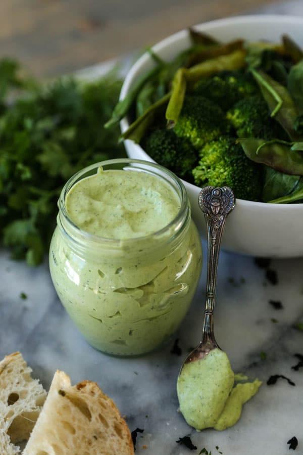 A jar of green goddess dressing with a small silver spoon of dressing leaning on a white bowl of salad, and two pieces of bread in front of it with a pile of parsley in the background