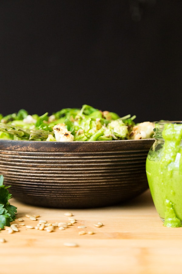 Green Spring Vegetable salad in a wood bowl on a wood surface with sunflower seeds sprinkled in front and part of a glass jar of creamy chimichurri sauce