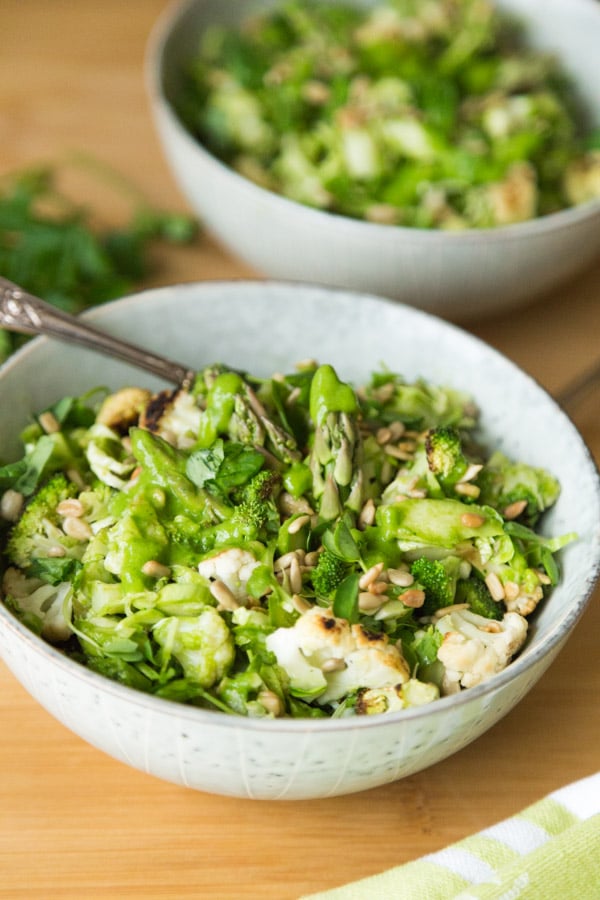 Two white bowls with green spring vegetable and creamy chimichurri sauce with a gold spoon in one and a green and white napkin in the front of the bowl