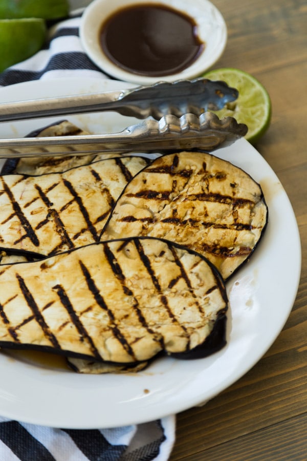 Grilled eggplant with lime zest on a white plate with tongs on a wood board