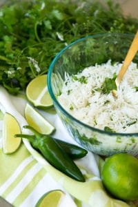 A glass bowl filled with cilantro lime rice on a green and white towel with a gold spoon in the bowl with jalapeno peppers, limes and a bunch of cilantro around it.