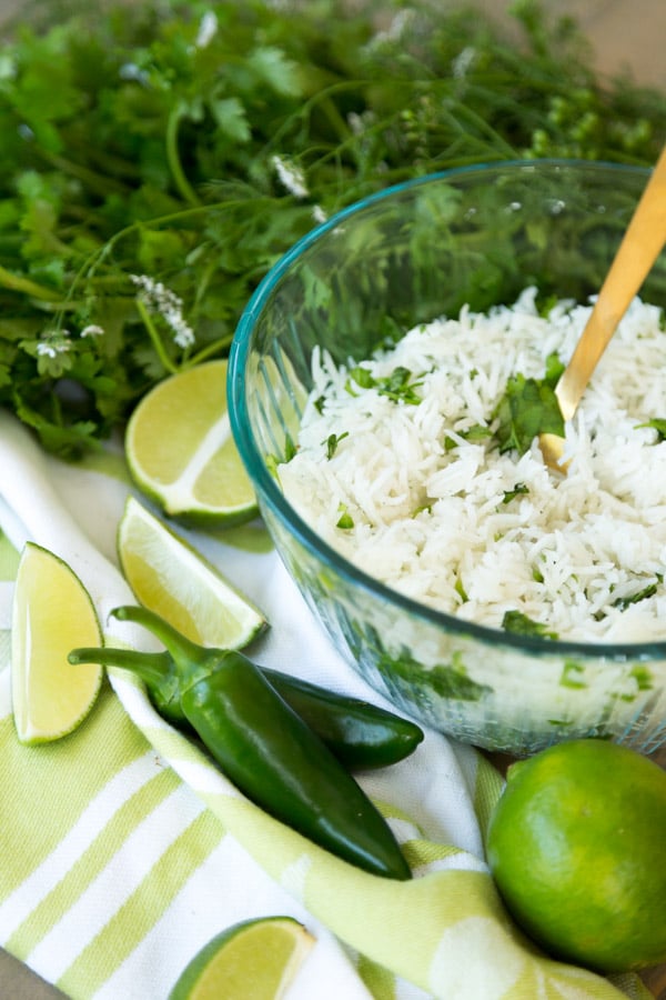 A glass bowl filled with cilantro lime rice on a green and white towel with a gold spoon in the bowl with jalapeno peppers, limes and a bunch of cilantro around it.