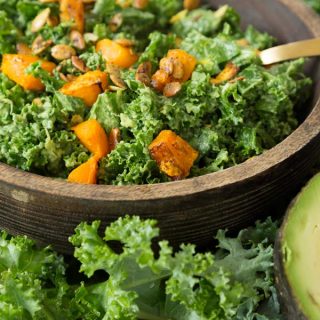 Kale and roasted squash salad in a wood bowl with a gold fork in the bowl and kale leaves and a sliver of avocado next to the bowl
