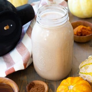 A mason jar filled with pumpkin spice cashew milk with brown wood bowls of spices and maple syrup next to it, and pumpkins surrounding the jar