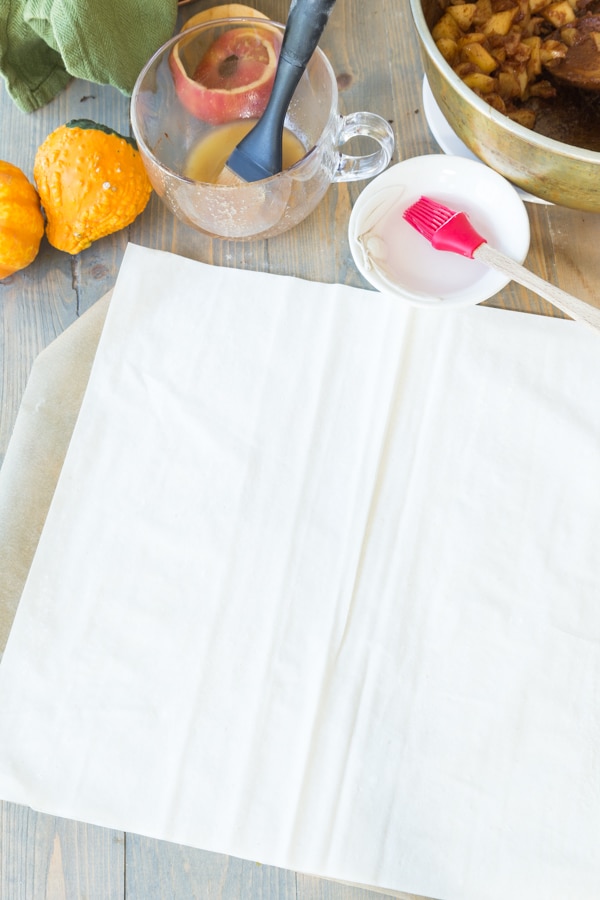 Two pieces of phyllo dough on parchment paper with a small white bowl of water and a red pastry brush next to it
