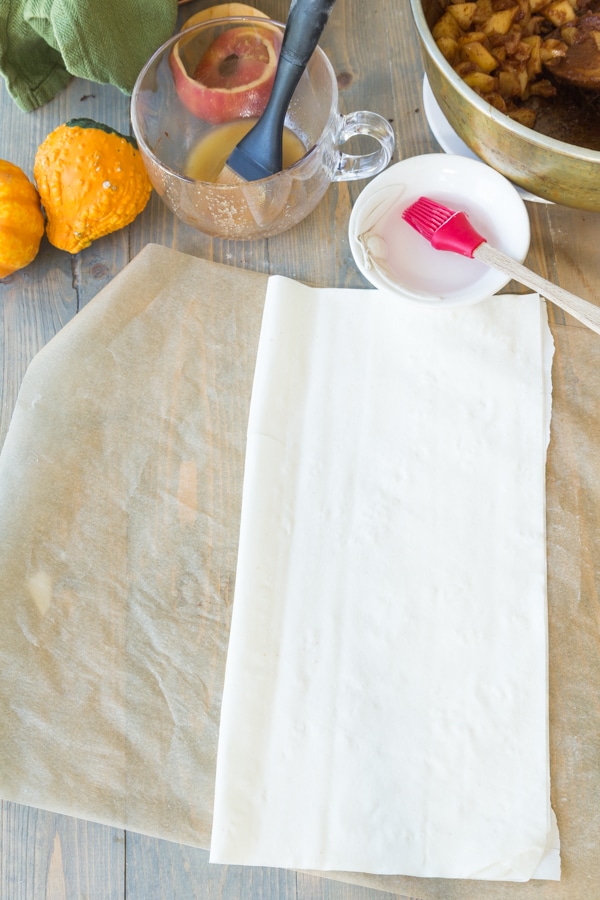 2 pieces of phyllo dough on parchment paper folded in half with pumpkins and bowls of melted butter and water in the background