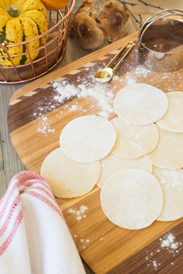 A floured wood board with a stack of raw wonton wrappers and a gold measuring spoon and a measuring cup, and a gold basket of squash in the background.