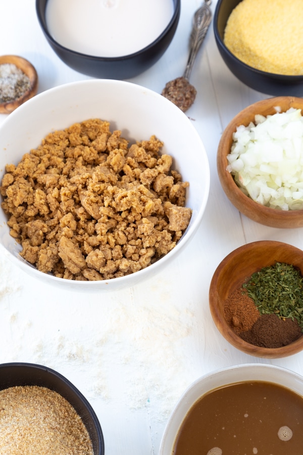 A white bowl with pulsed soy curls in it, and several wood and black bowls with chopped onion, spices, and ingredients for Swedish meatballs.
