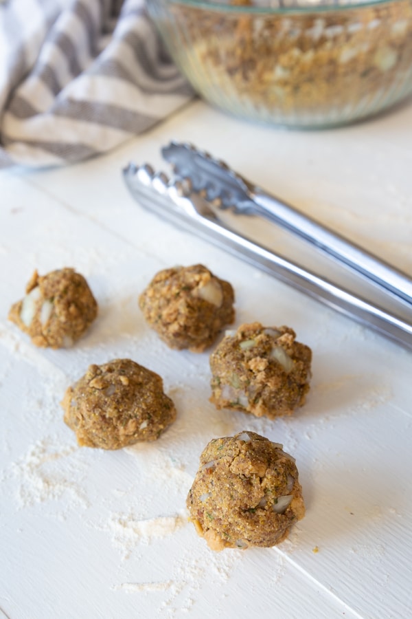 Uncooked vegan Swedish meatballs on a white board with silver tongs and a blue and white dish towel next to them